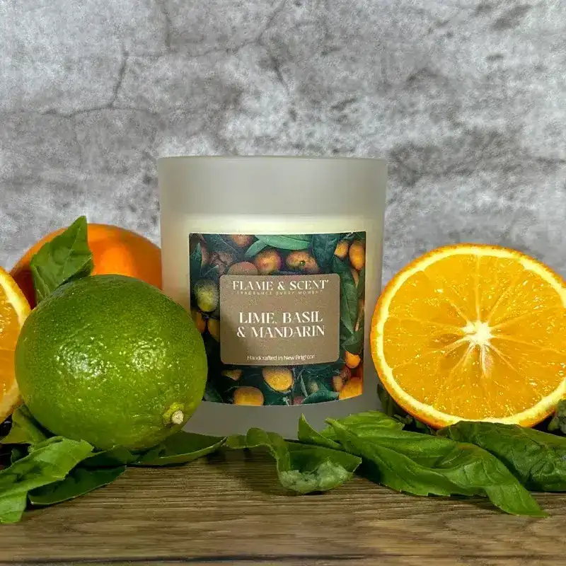 Flame & Scent The Lime, Basil & Mandarin Candle sits on a wooden surface with fresh basil leaves, a whole lime, an orange, and a sliced orange half, set against a grey textured background.