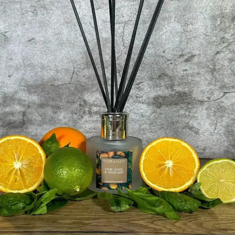 Flame & Scent The Lime, Basil & Mandarin Reed Diffuser with black reeds rests on a wooden surface surrounded by fresh lime, orange and lemon halves, plus basil leaves, set against a grey textured background.