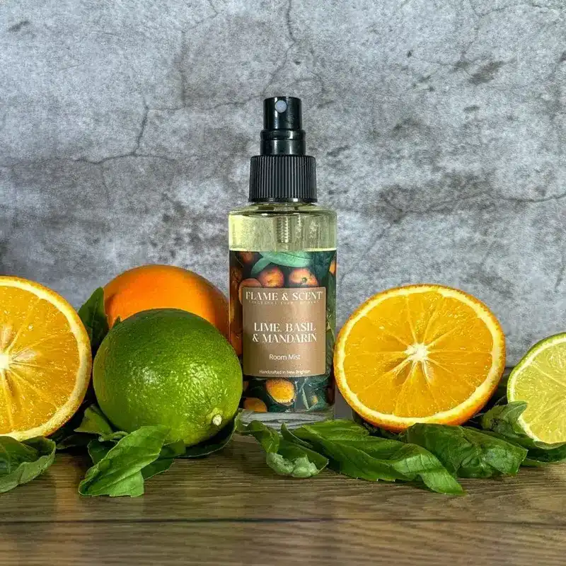 Flame & Scent The Lime, Basil & Mandarin Room Mist is displayed with fresh basil leaves, mandarin slices, a lime, and oranges on a grey textured background.