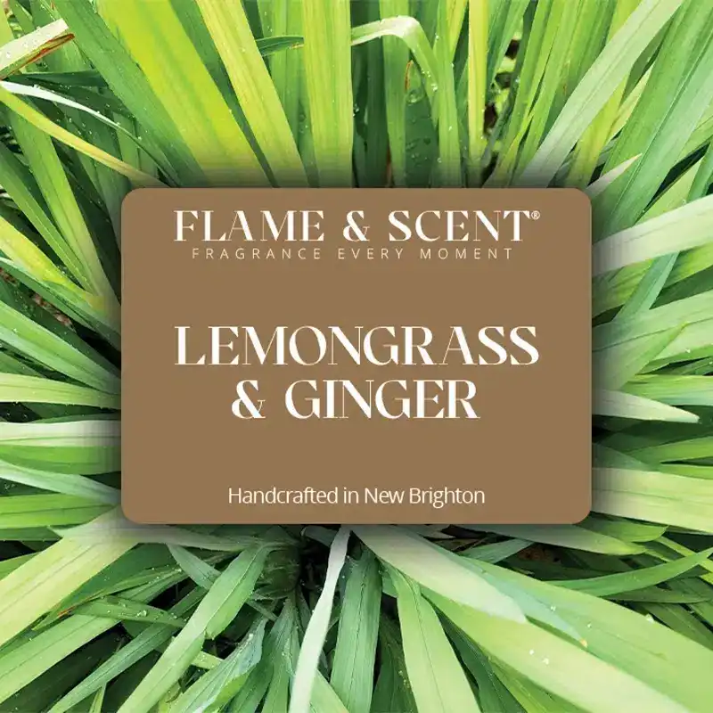 Flame & Scent A brown label with white text reads FLAME & SCENT FRAGRANCE EVERY MOMENT Lemongrass & Ginger Candle Handcrafted in New Brighton, set over green lemongrass leaves—perfect for a refreshing citrus or spicy ginger aroma.