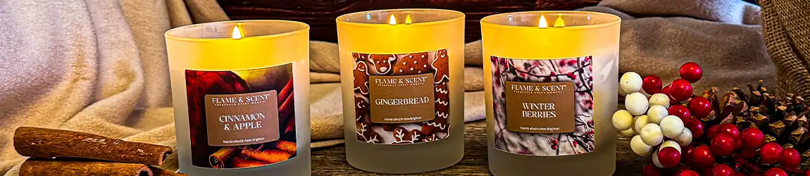 Christmas Banner Three lit scented candles labelled Cinnamon & Apple, Gingerbread, and Winter Berries sit on a cosy surface surrounded by cinnamon sticks, pinecones, red-and-white berries, and soft fabric. Flame & Scent