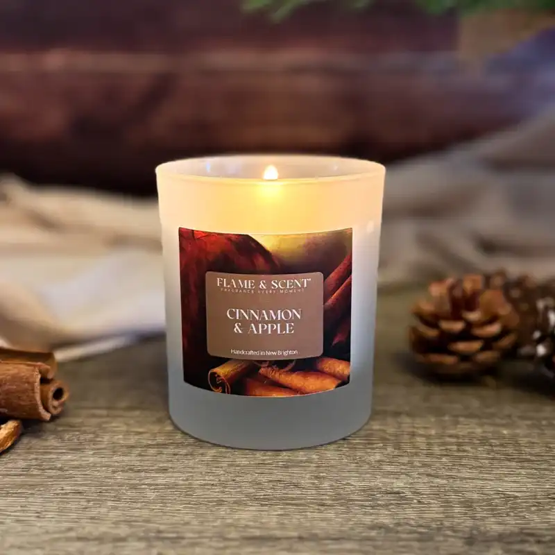 A lit Cinnamon & Apple Candle in a frosted glass jar rests on a wooden surface, surrounded by cinnamon sticks, a pine cone, and a softly blurred cloth in the background. Flame & Scent