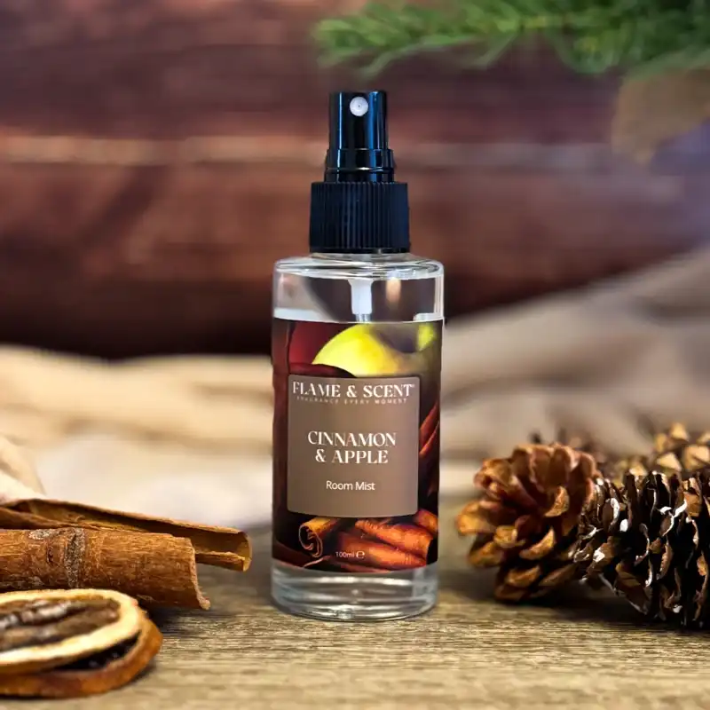 The Cinnamon & Apple Room Mist rests on wood, surrounded by pine cones, dried orange slices, a cinnamon stick, and evergreen branches, filling the air with warm cinnamon and apple fragrance for a cosy holiday feel. Flame & Scent