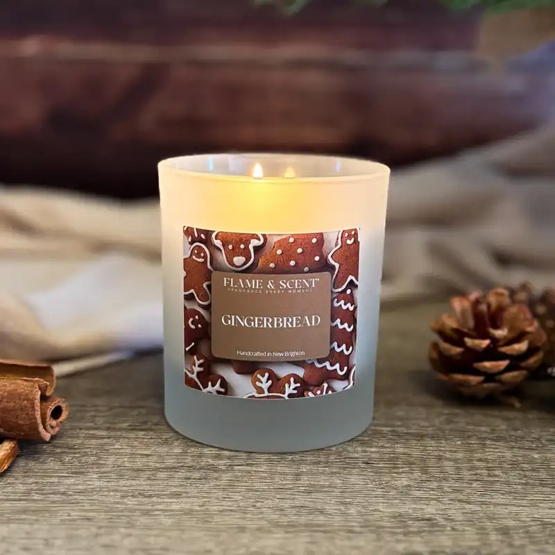 A Cinnamon & Apple Candle in a frosted glass holder sits on a wooden surface near pinecones and cinnamon sticks. The label shows gingerbread biscuits, creating an inviting ambience with its cosy scented glow. Flame & Scent