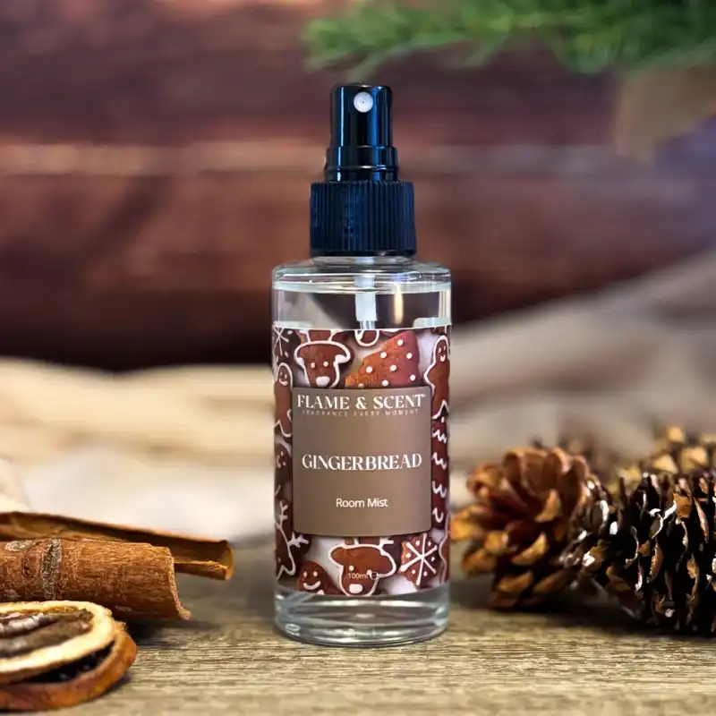 A bottle of Cinnamon & Apple Room Mist with a festive label sits on a table beside pine cones, dried orange slices, an apple, and a cinnamon stick, with blurred greenery and a wooden background. Flame & Scent