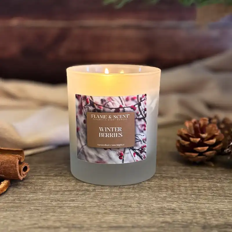 A lit Cinnamon & Apple Candle sits on a wooden surface, surrounded by a pine cone, cinnamon sticks, and a neutral cloth, with a blurred rustic background. Flame & Scent
