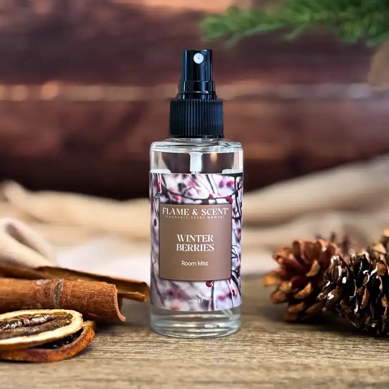 A clear spray bottle labelled Cinnamon & Apple Room Mist sits on a wooden surface with cinnamon sticks, dried orange slices, and pine cones nearby. A pine branch and blurred wooden background create a cosy vibe, evoking scents of cinnamon and apple. Flame & Scent